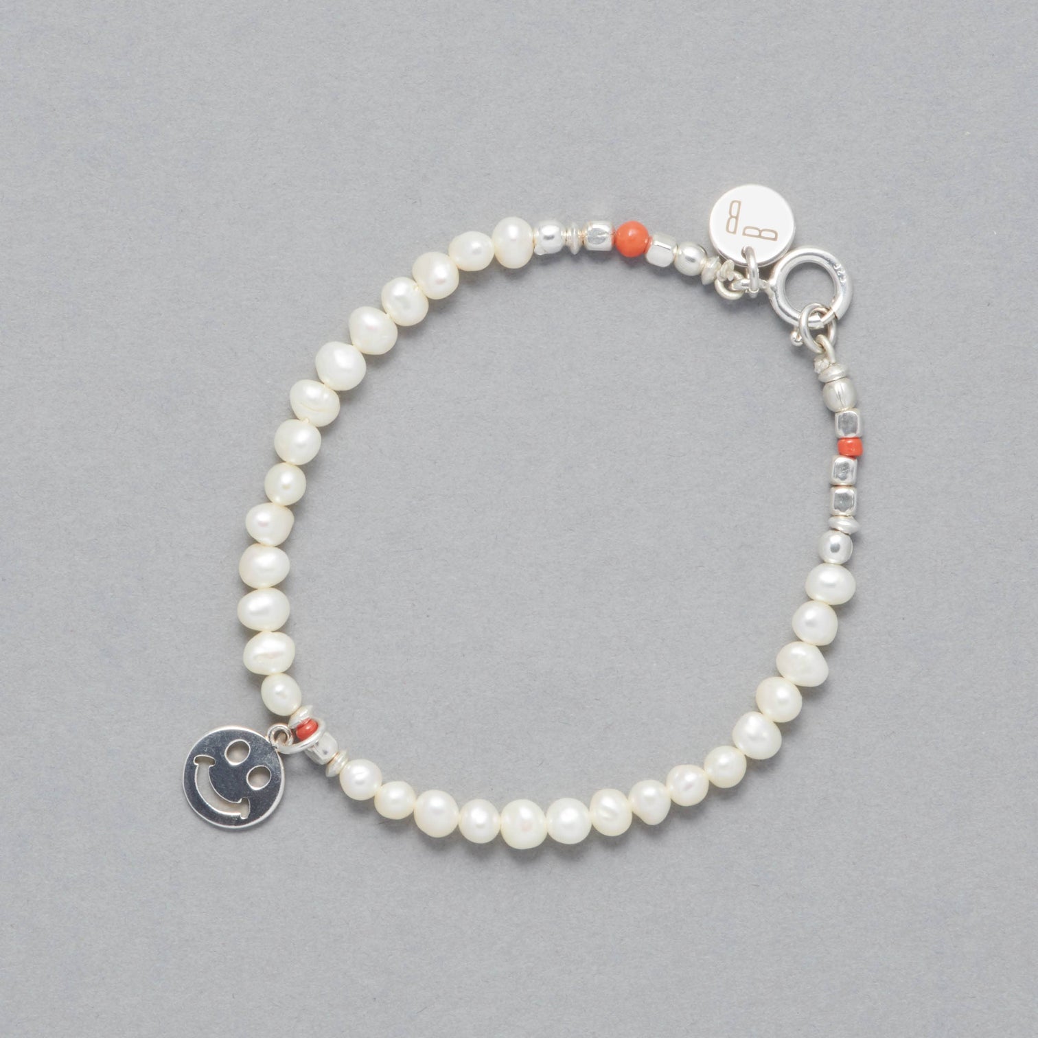 The ALAIA Bracelet is made with cultured freshwater white pearls, a sterling silver smiley charm and elements and a touch of coral.