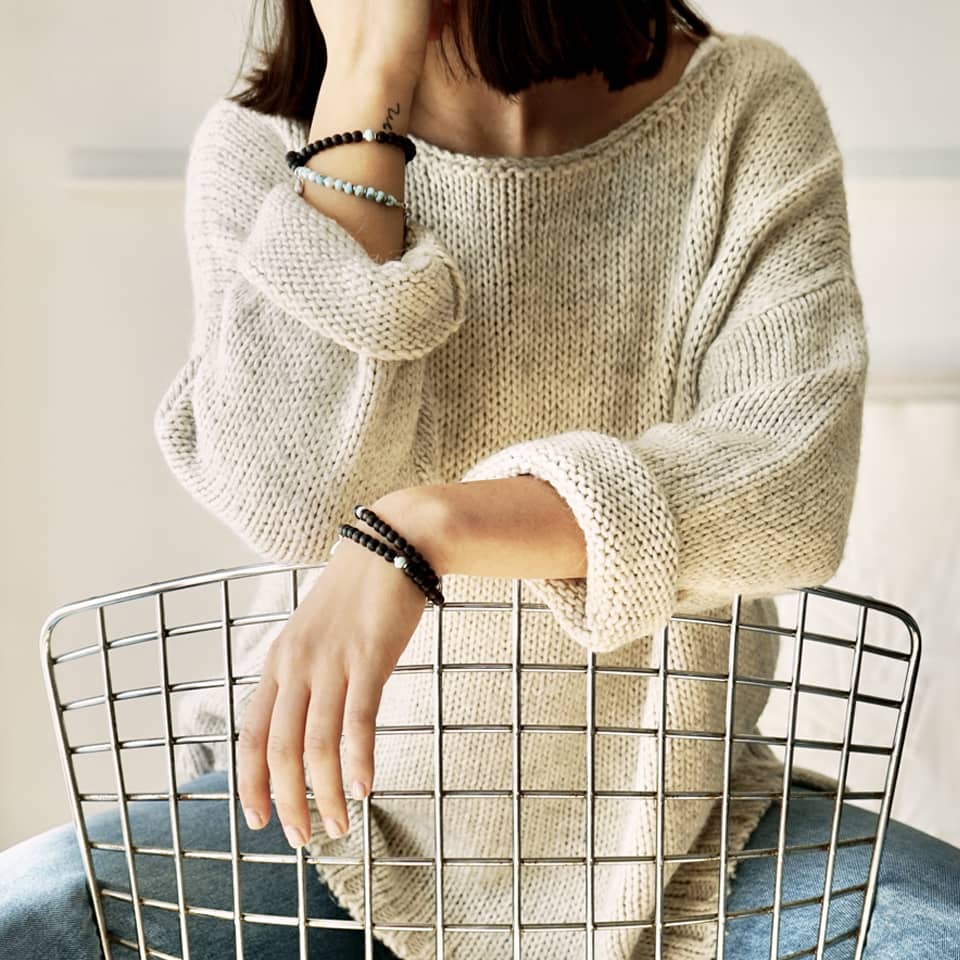 Picture of a female model sitting on a chair. Only the upper part of the body without the face is visible. She is wearing on both wrists the LE BIJOUBIJOU KAIA Double Wrap Bracelets made with Larimar and Ebony. 