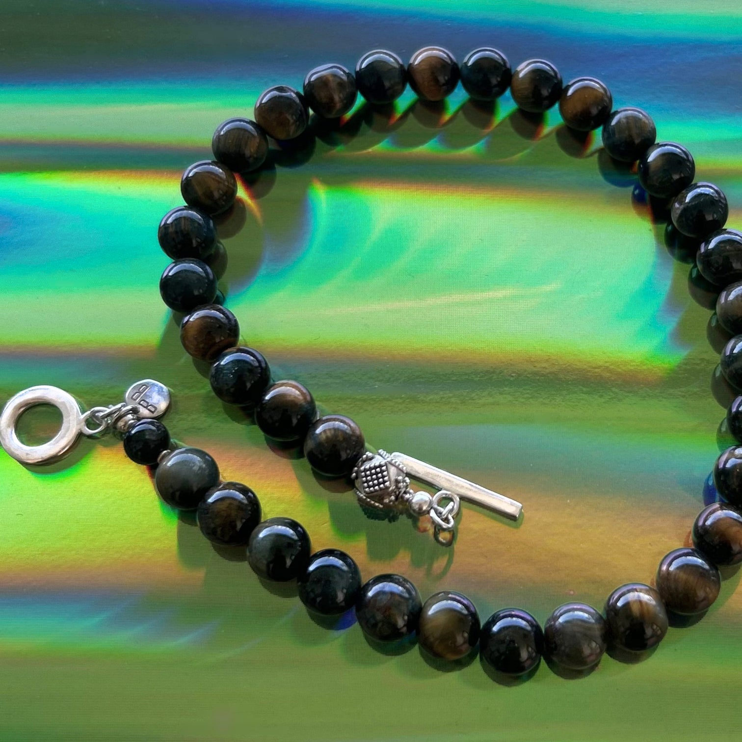 Amber Flash Necklace with round Tiger Eye beads and  an accent of a beautifully crafted silver bead next to a silver toggle clasp on a rainbow-reflective surface — Le Bijou Bijou” 