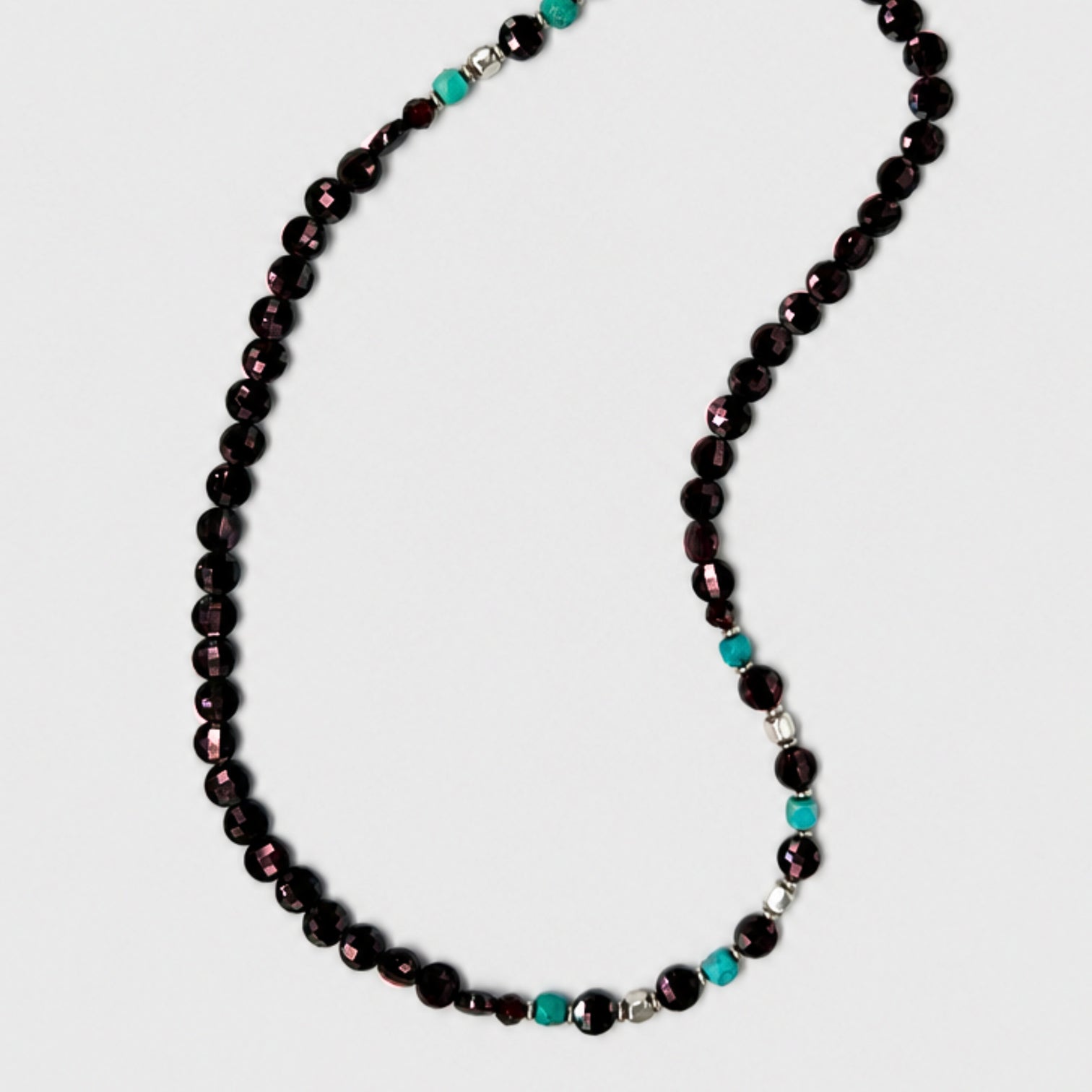 A beaded necklace featuring dark, faceted garnet rondelles with accents of square diamond-polish turquoises and silverl beads, displayed on a light gray background.