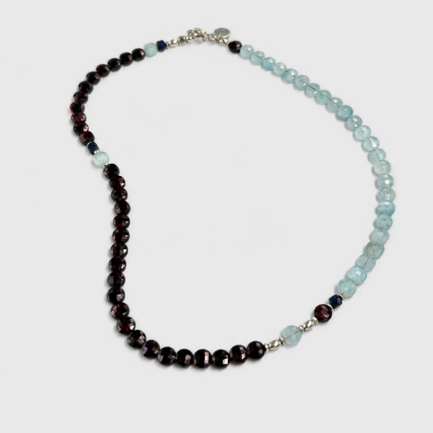 A beaded necklace featuring light blue faceted aquamarine rondelles on one side and dark red faceted rondelles on the other side, separated by small silver spacer beads and a single light blue bead. Clasp with the LeBijouBijou charm shown on a light gray background.