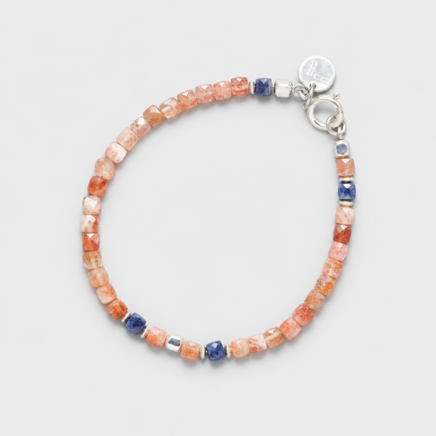 GINGER bracelet with faceted peach sunstones, blue sapphires, sterling silver clasp and round charm on a neutral light-grey background.