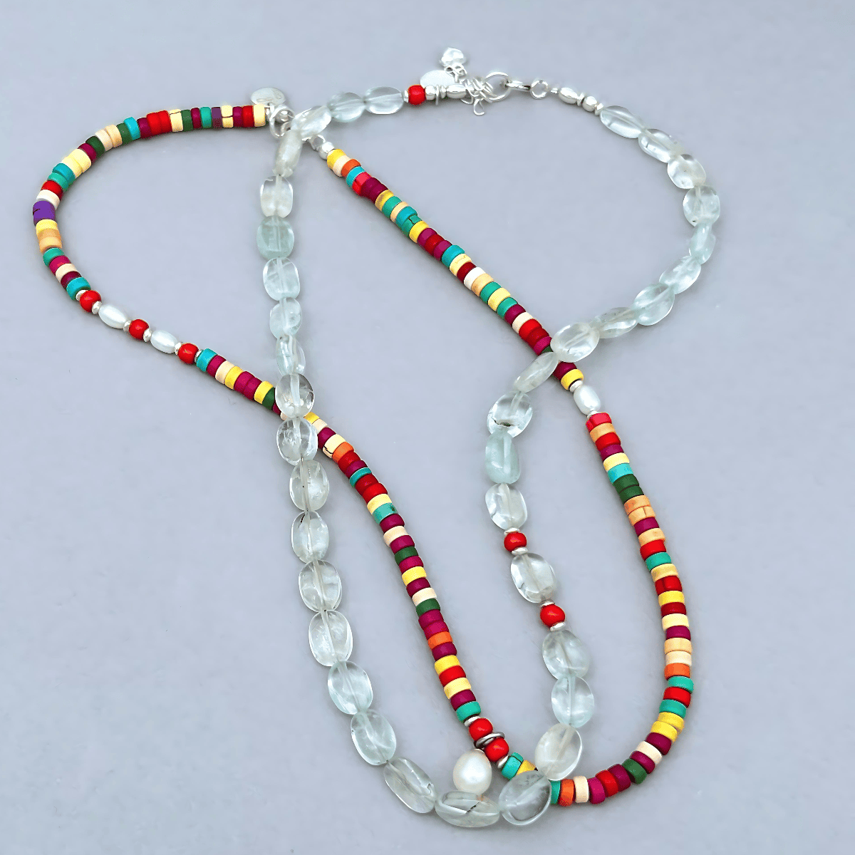 A beaded necklace featuring translucent light blue Aquamarine oval beads, combined with colourful cylindrical beads in red, yellow, green, and orange, displayed on a neutral gray background.