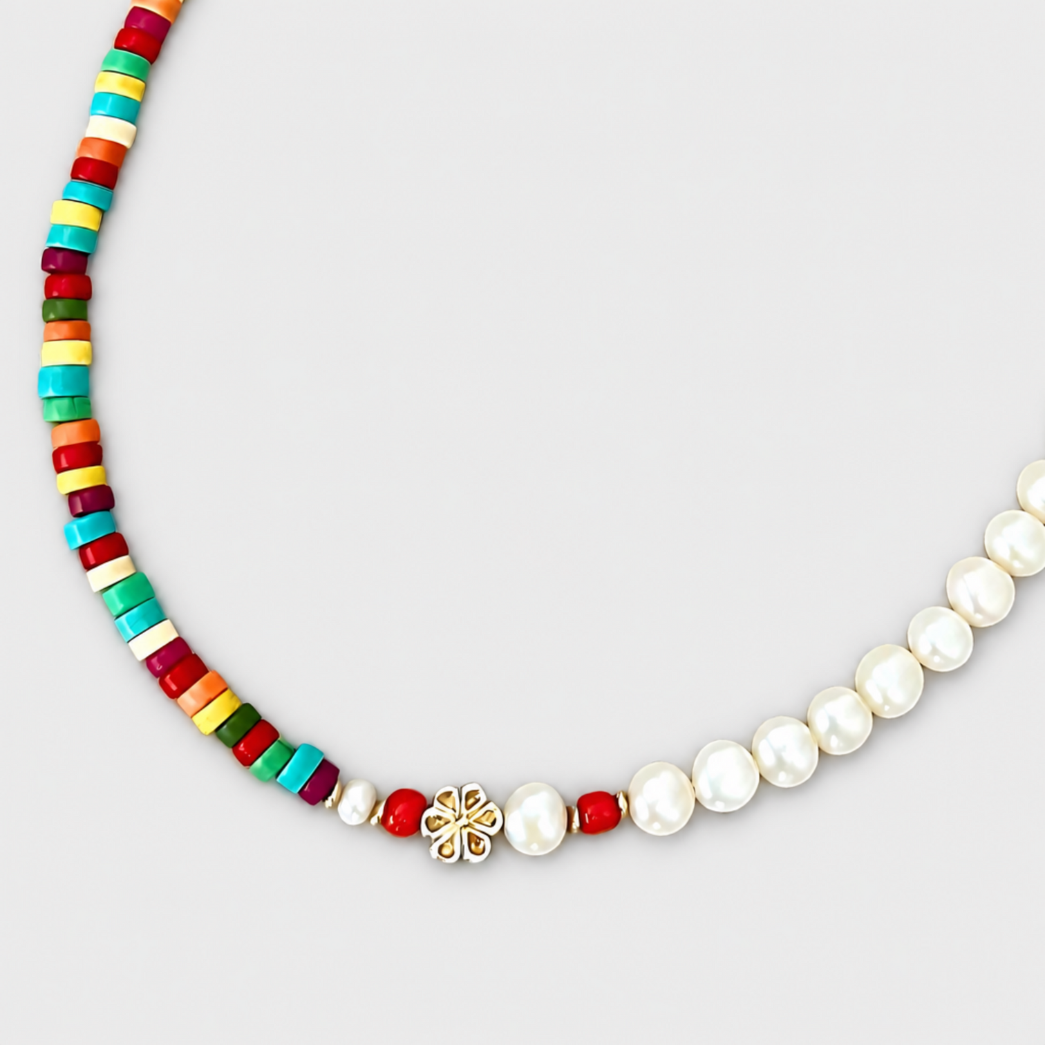 Close-up of HAPPY necklace showing multicoloured discs, white pearls, corald beads and a silver flower detail on a light grey background.