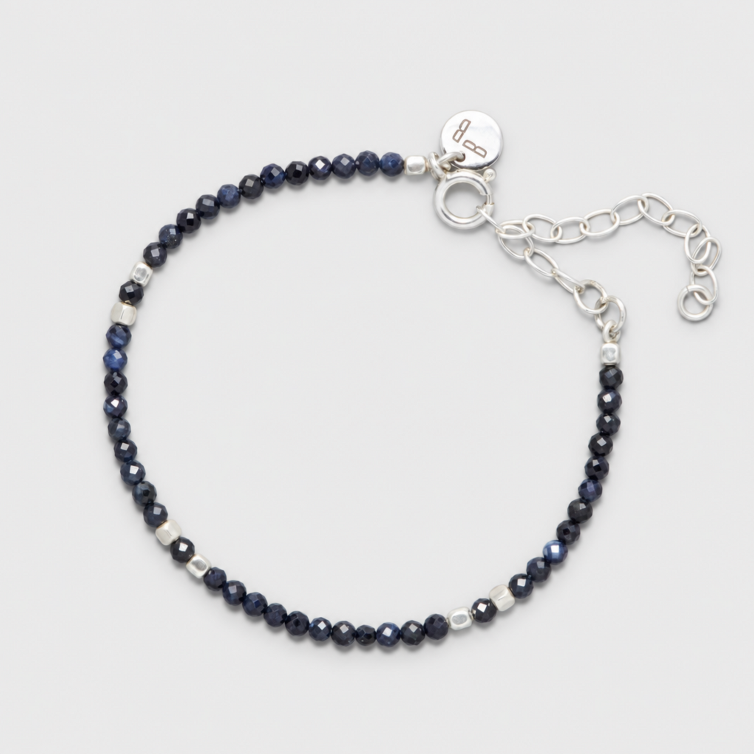 INDIGO anklet with faceted dark blue sapphires, sterling silver spacers, clasp, round charm and extension chain on a neutral light-grey background.