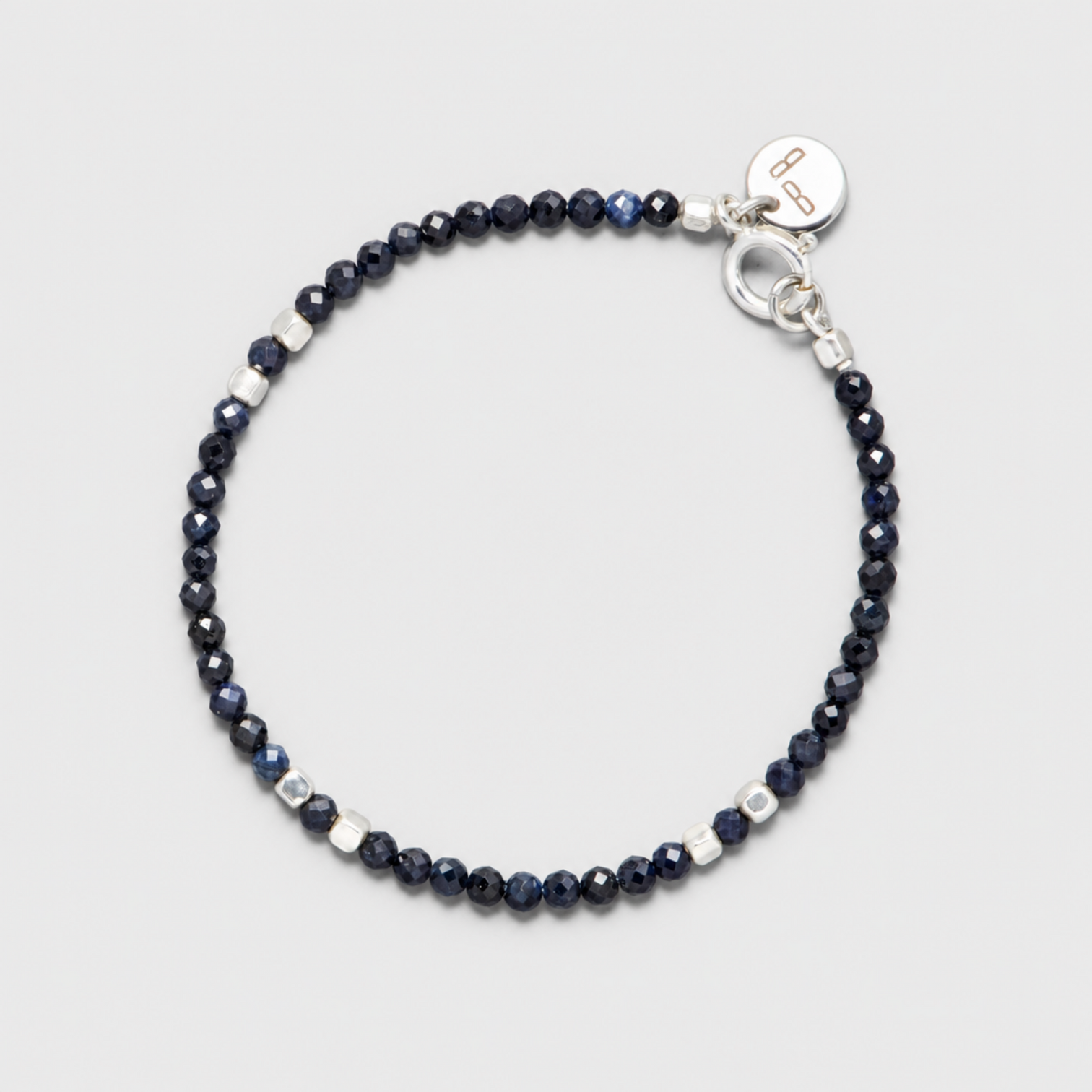 INDIGO bracelet with faceted dark blue sapphires, sterling silver spacers, clasp and round charm on a neutral light-grey background.