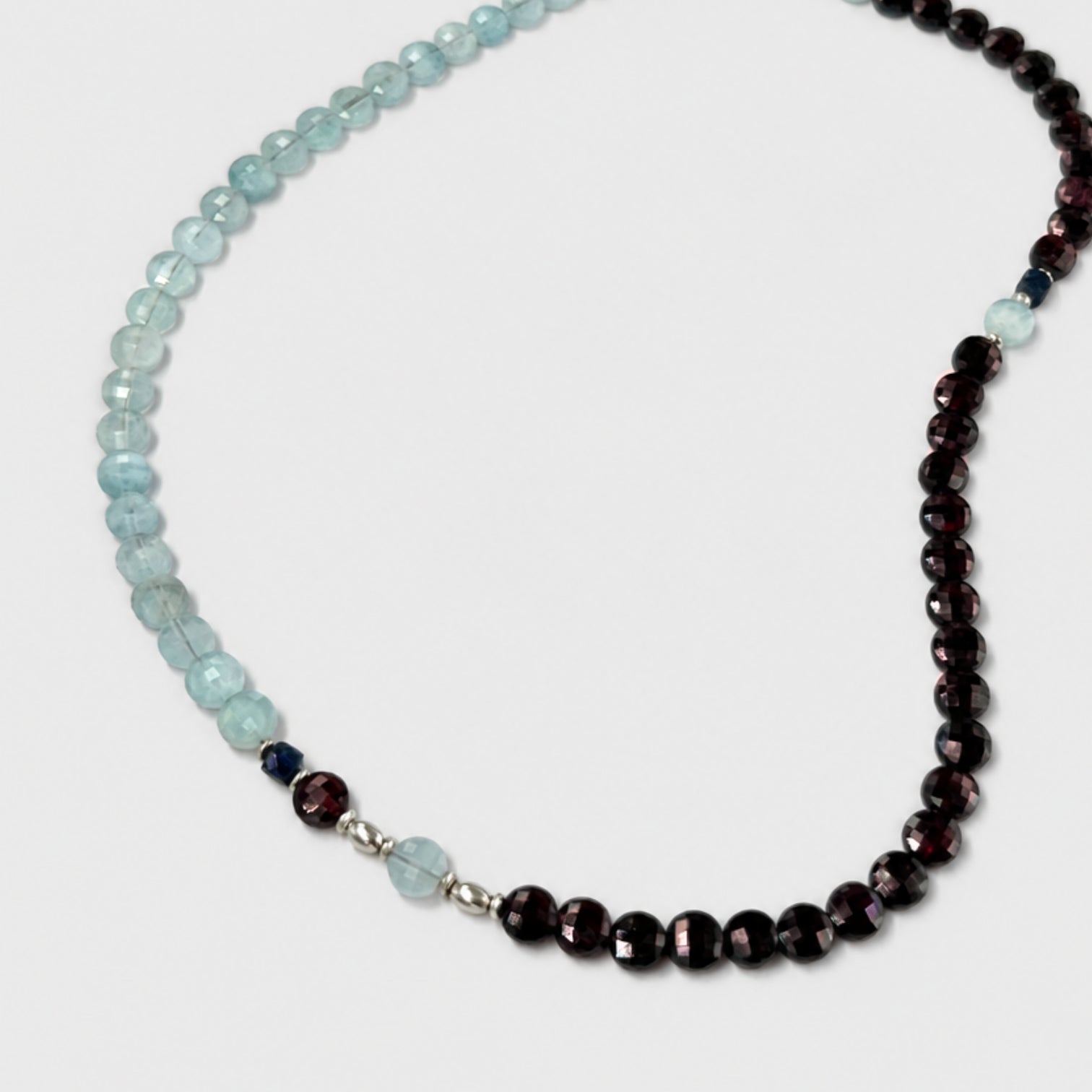 A beaded necklace featuring light blue faceted aquamarine rondelles on one side and dark red faceted rondelles on the other side, separated by small silver spacer beads and a single light blue bead, shown on a light gray background.