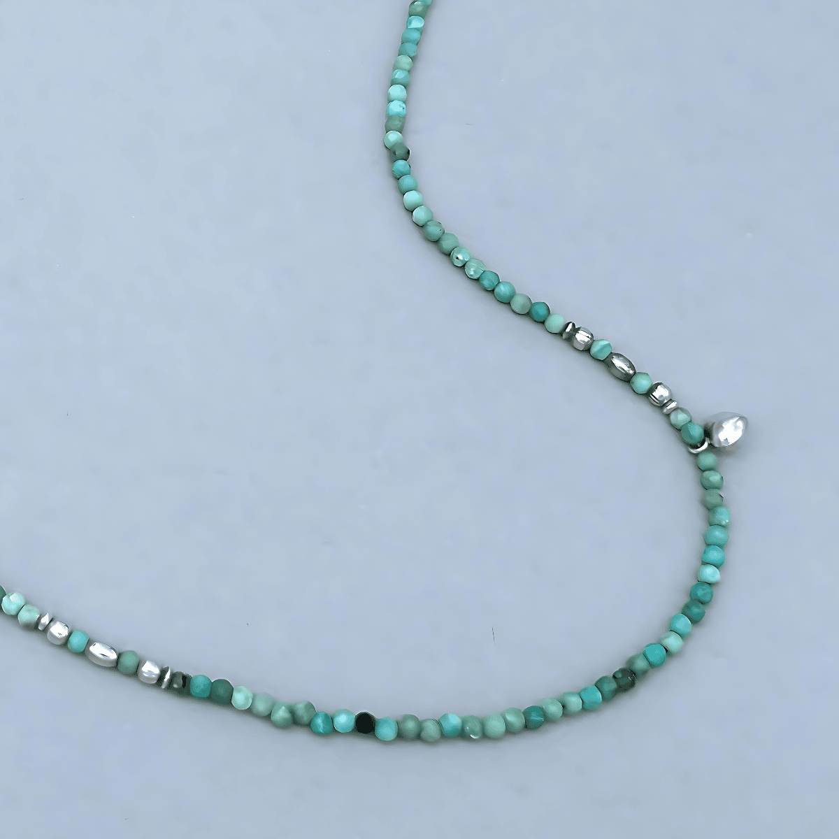 Detail of the Le BijouBijou JOY Necklace showing the turquoise faceted Opal beads, sterling silver spacer elements and a small silver heart charm on a light grey background.

