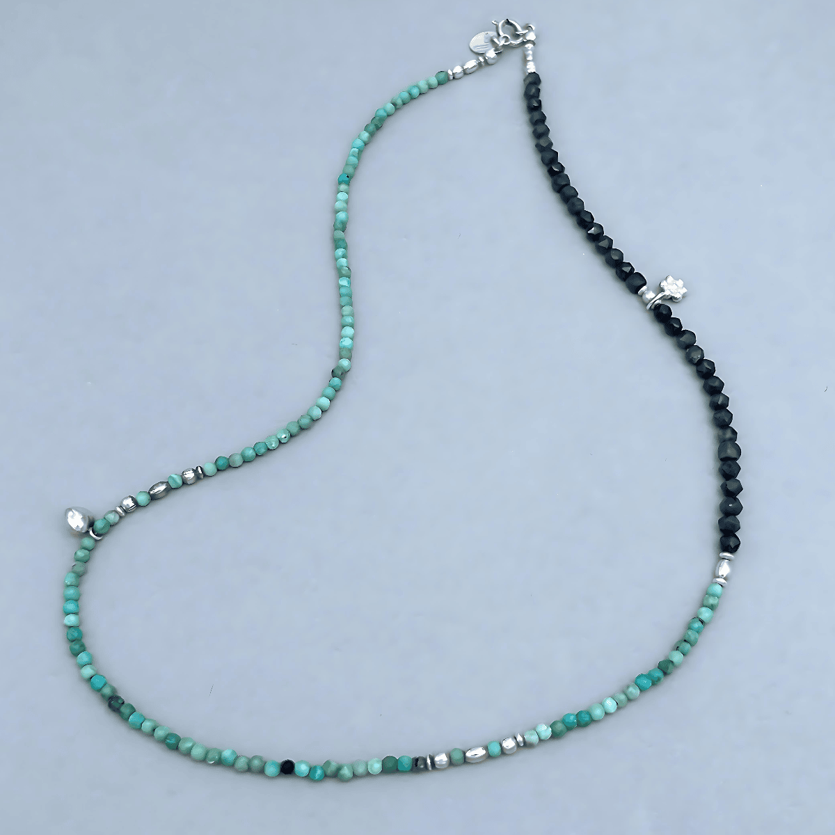 Le BijouBijou JOY Necklace laid flat on a light blue-grey surface, showing the full length with bluish-turquoise faceted Opal beads, dark navy Sapphire cubes, sterling silver spacers and two small silver charms.