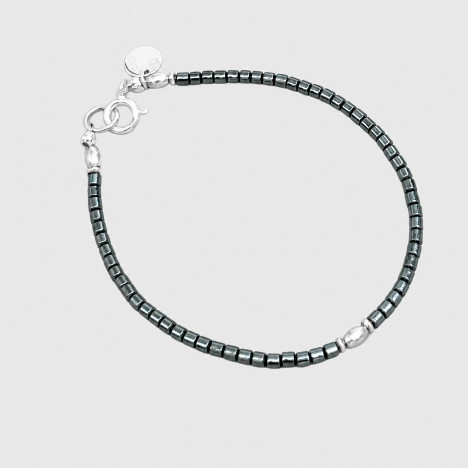 MIDNIGHT SILVER bracelet with dark metallic cylindrical beads, an oval silver accent, silver clasp and round charm on a neutral light-grey background.