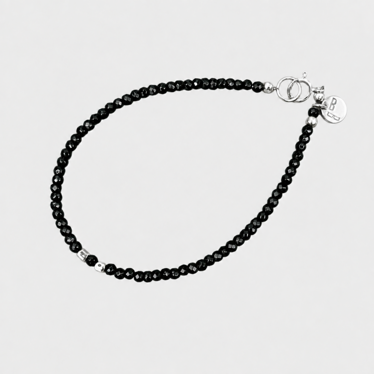 MIDNIGHT SPARKLE bracelet with faceted black onyx beads, silver spacers, clasp and round logo charm on a neutral light-grey background.