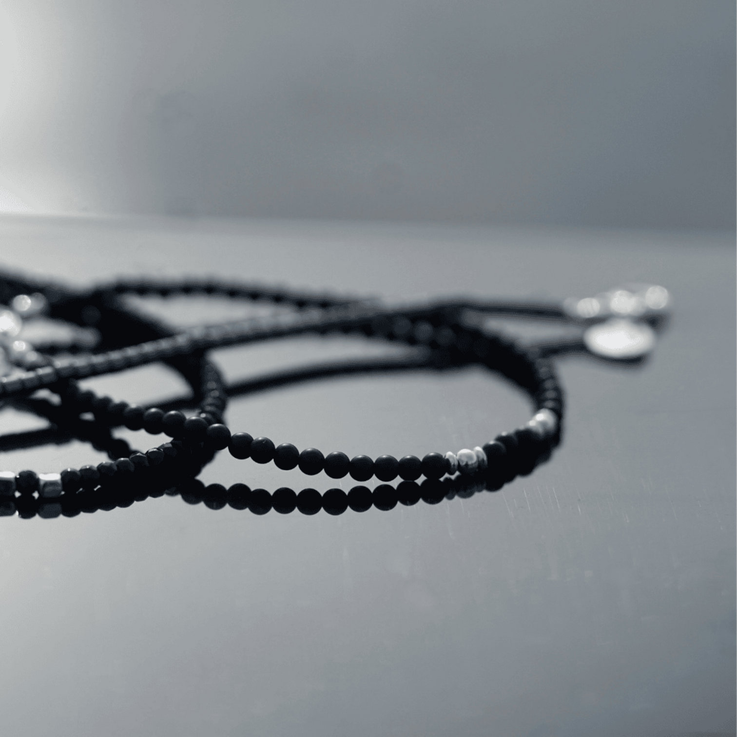 A close-up of a beaded necklace with small black beads and a few silver spacer beads, resting on a reflective gray surface.