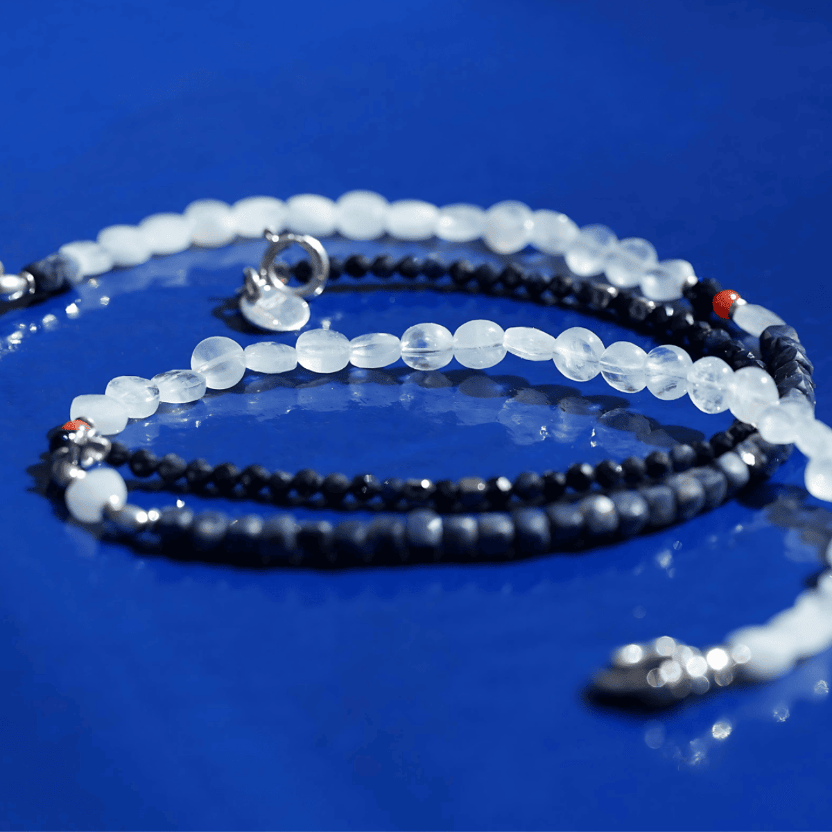 Le BijouBijou Perfect Match Necklace coiled on a glossy blue surface, showing pale Aquamarine rounded beads, dark faceted Sapphire beads, red Coral accents and a sterling silver clasp.