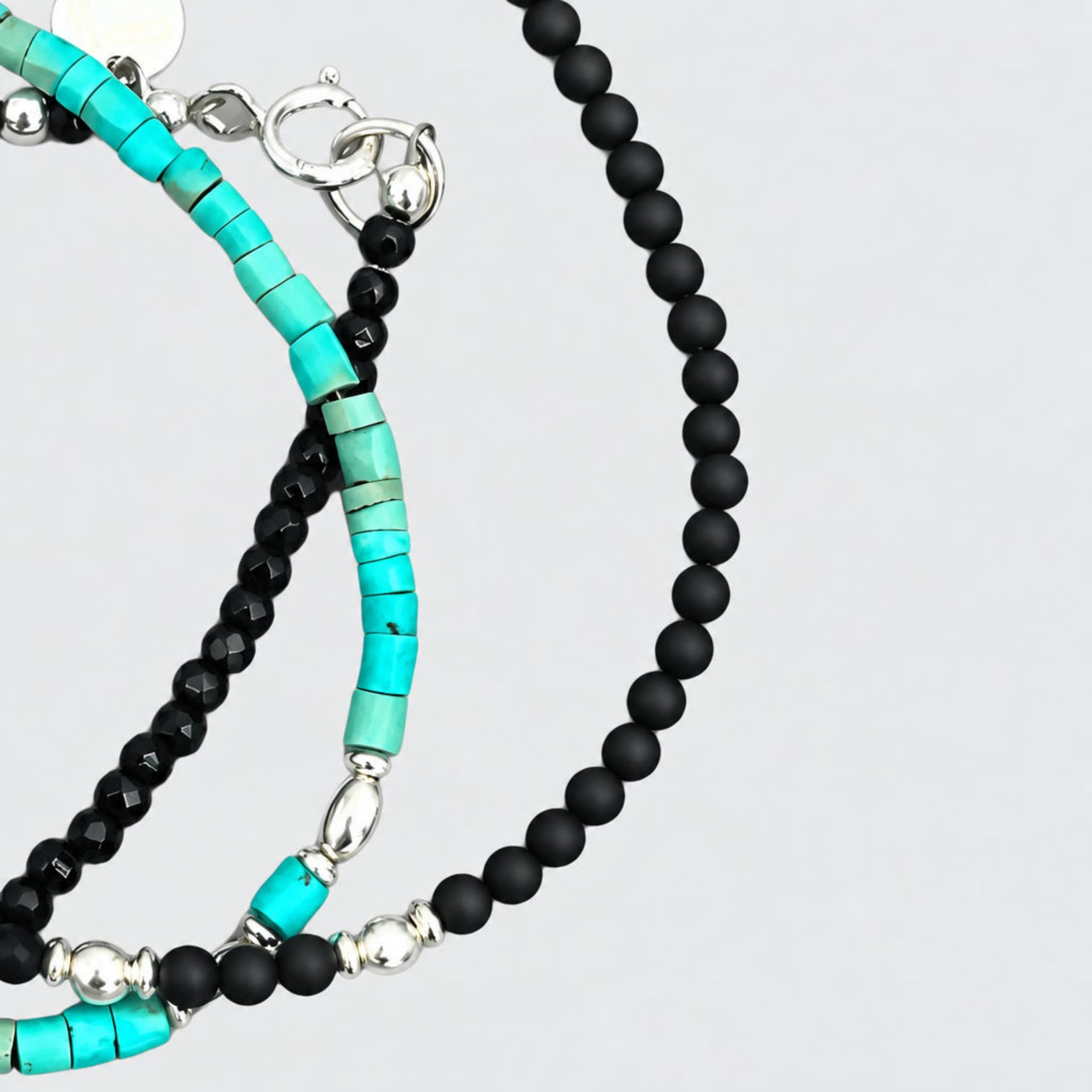 Detail of the MAGICIAN matte black bead bracelet with silver spacers, alongside shiny black and turquoise bead bracelets with silver clasps and round charms on a light-grey 