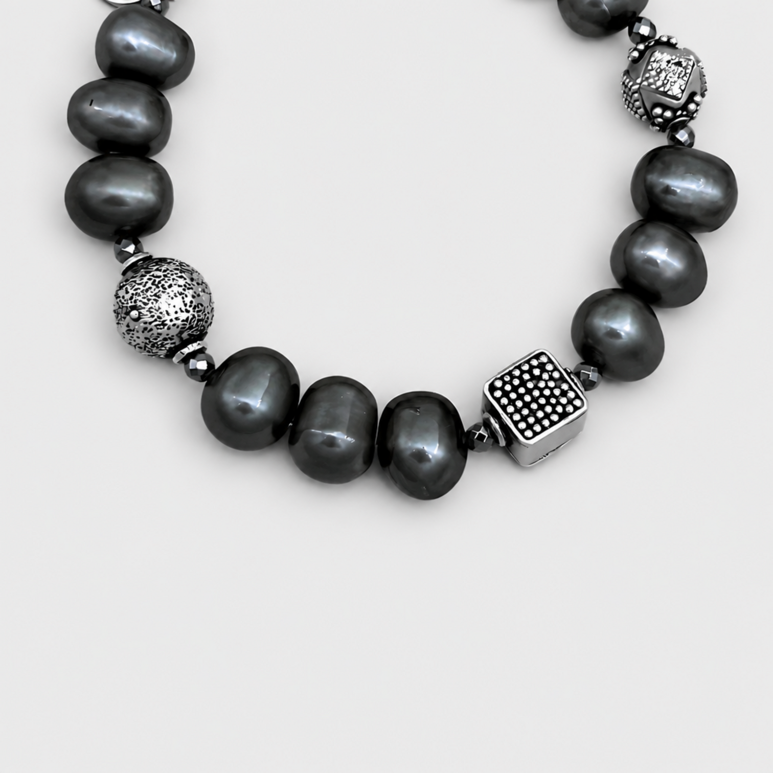 Close-up of NAIDA dark pearls with textured round and patterned square silver-tone beads on a light grey background.