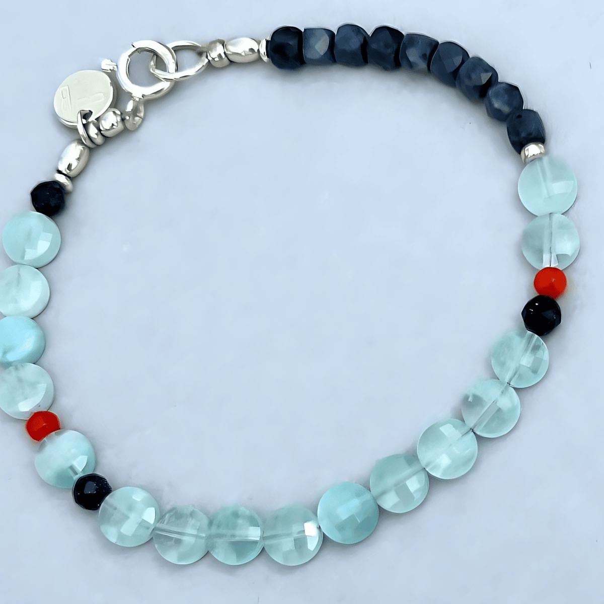 A beaded bracelet featuring faceted light blue aquamarine beads, dark navy blue Saphire beads, and tiny red accent of coral, completed with a silver clasp.