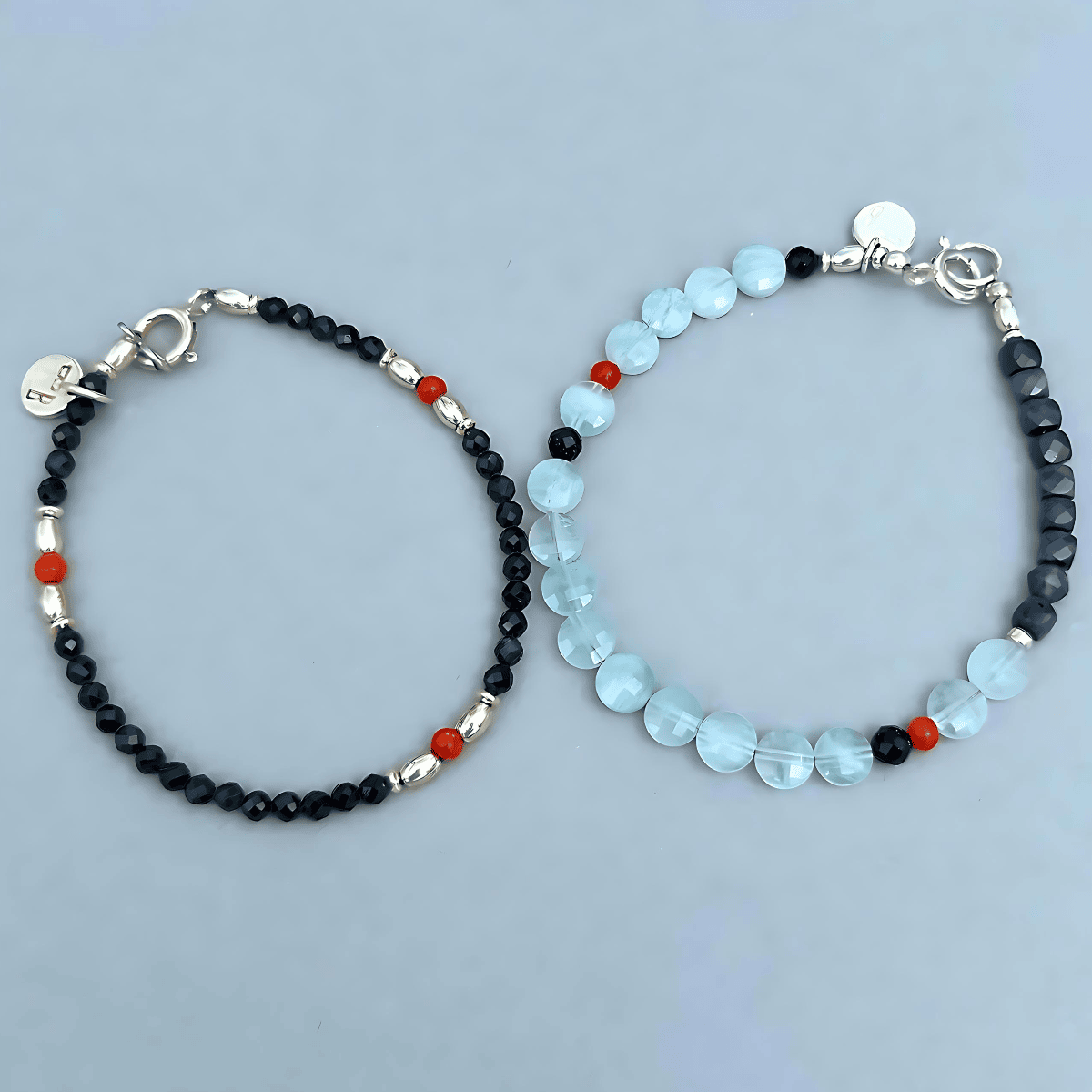 Two beaded bracelets displayed on a light blue-gray background. One bracelet features small dark blue Saphire beads with silver spacers and red coral accent beads.

