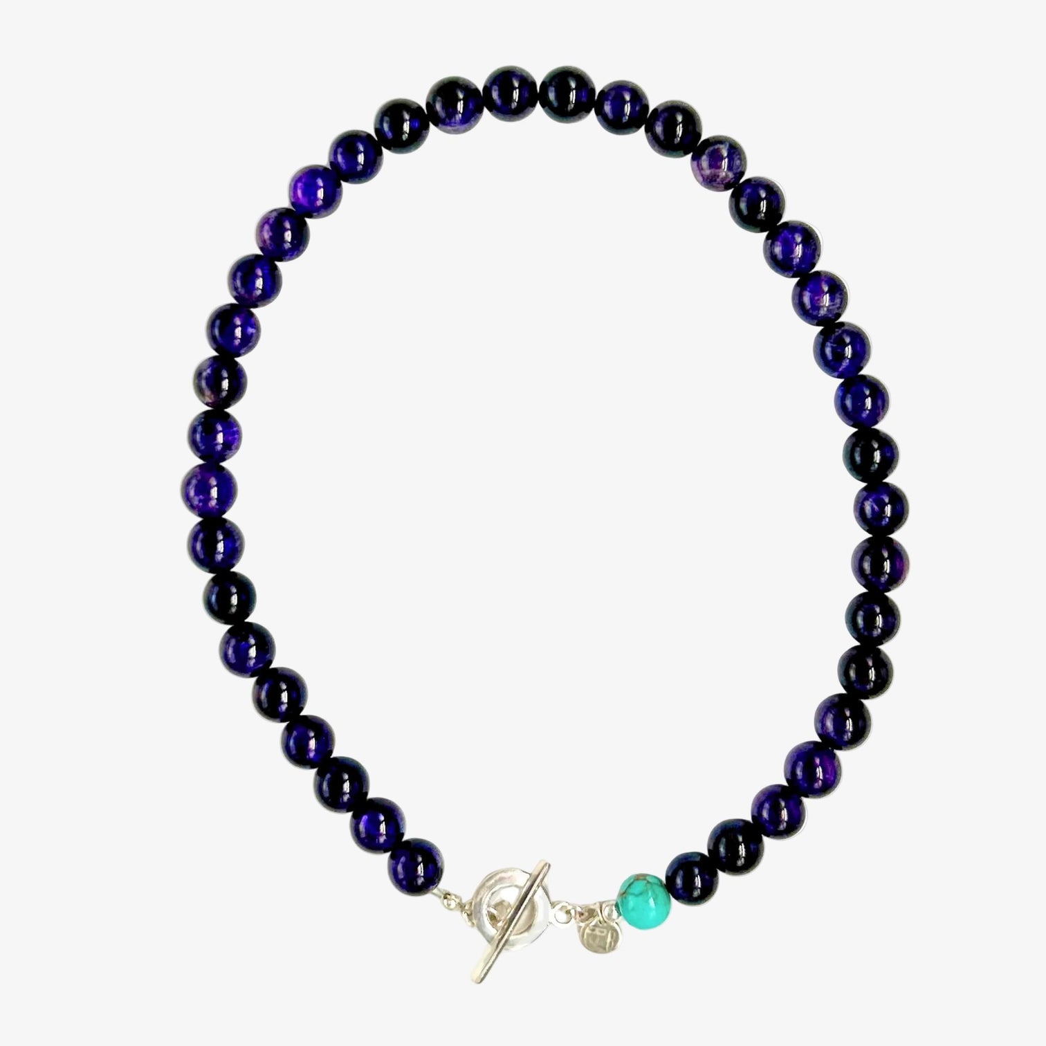 A beaded necklace made of glossy, dark purple amethyst round beads, with a silver-toned toggle clasp and a turquoise bead accent next to the clasp, is displayed on a neutral light grey surface.