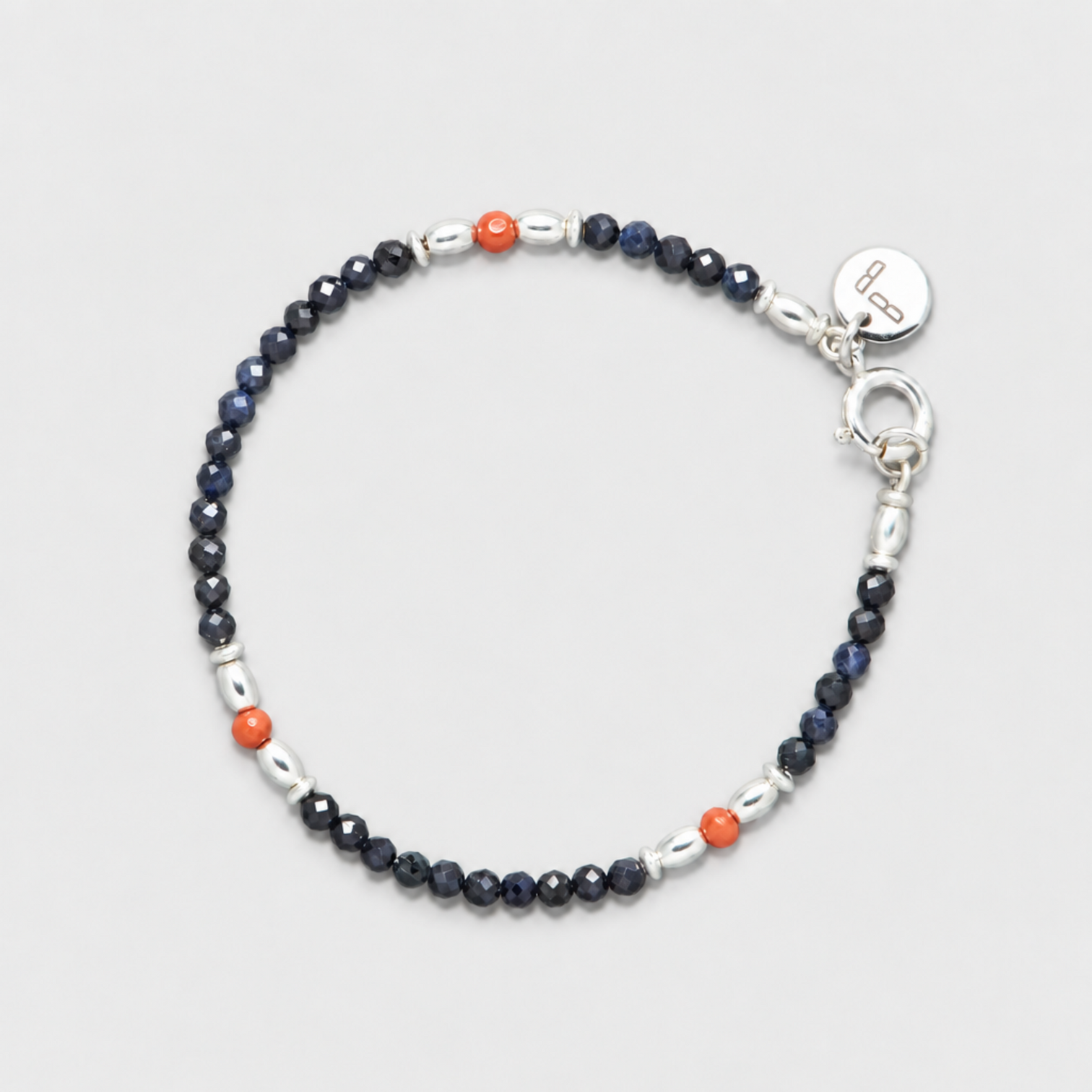 SKY bracelet with faceted dark blue sapphires, three red coral beads, sterling silver elements, clasp and round charm on a neutral light-grey background.