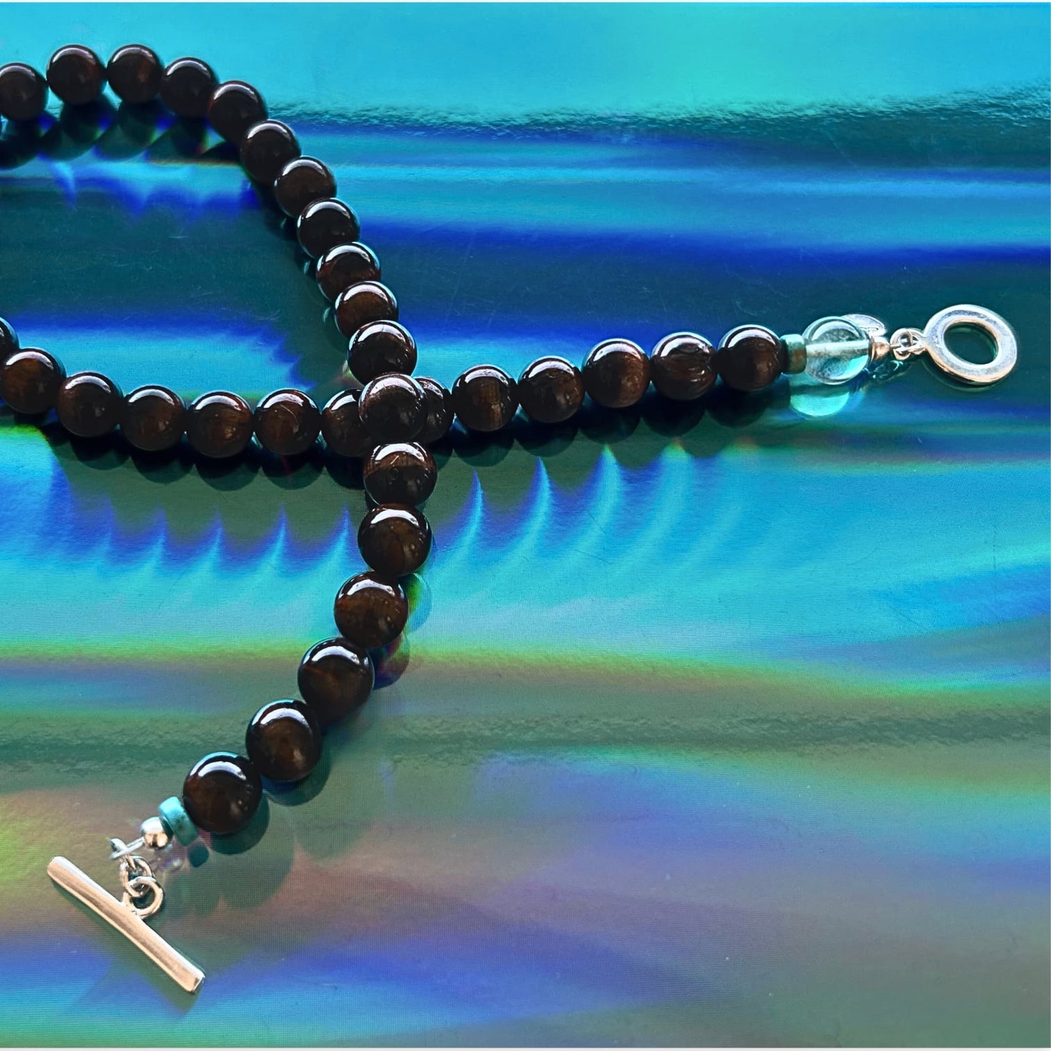 A beaded necklace of polished red-brown tiger's eye beads, with a silver toggle clasp and a fluorite bead with a silver logo charm, displayed on a vibrant, iridescent blue surface.