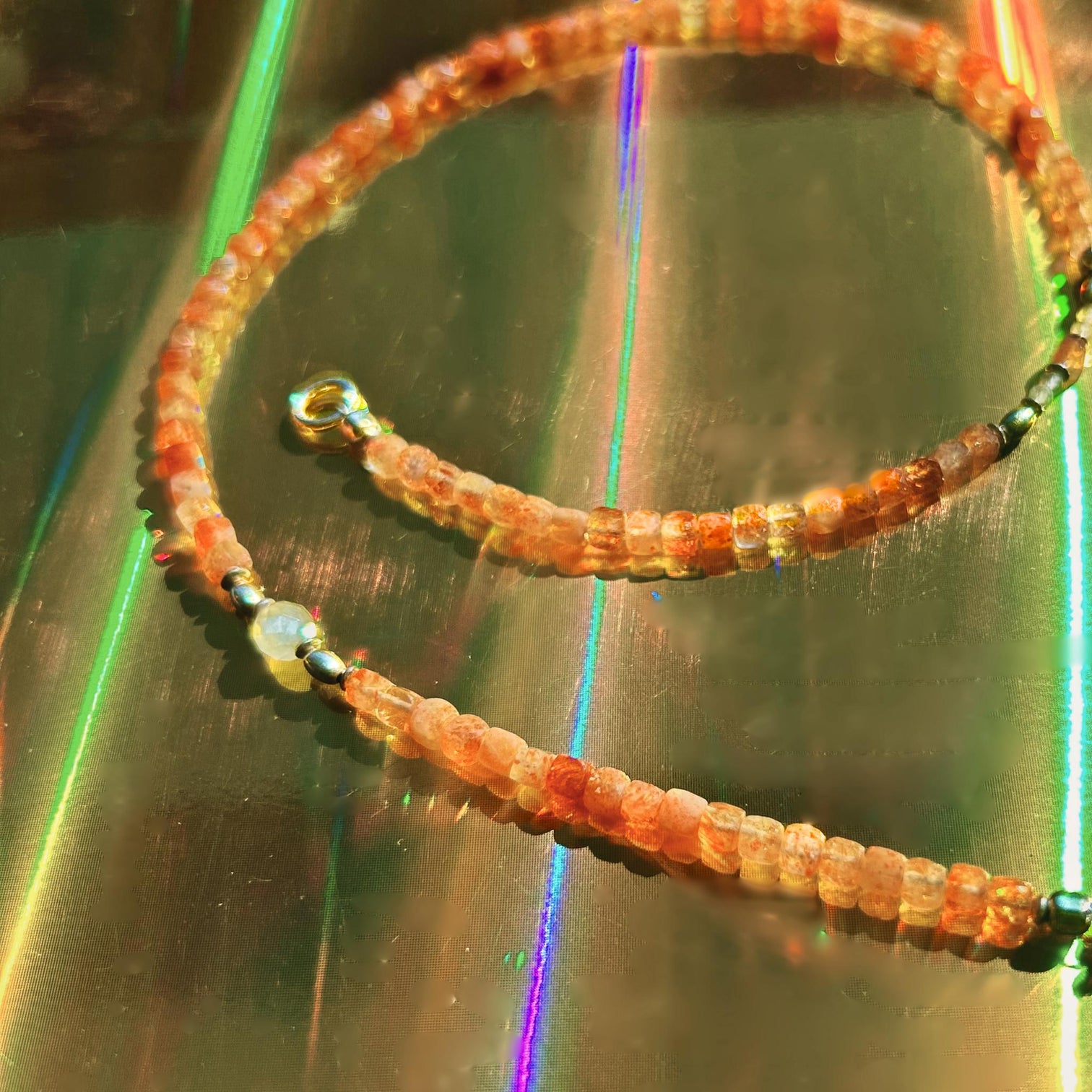 A beaded necklace with square faceted sunstone beads, one faceted moonstone bead, and silver spacer beads, displayed on a reflective surface with colourful light reflections.
