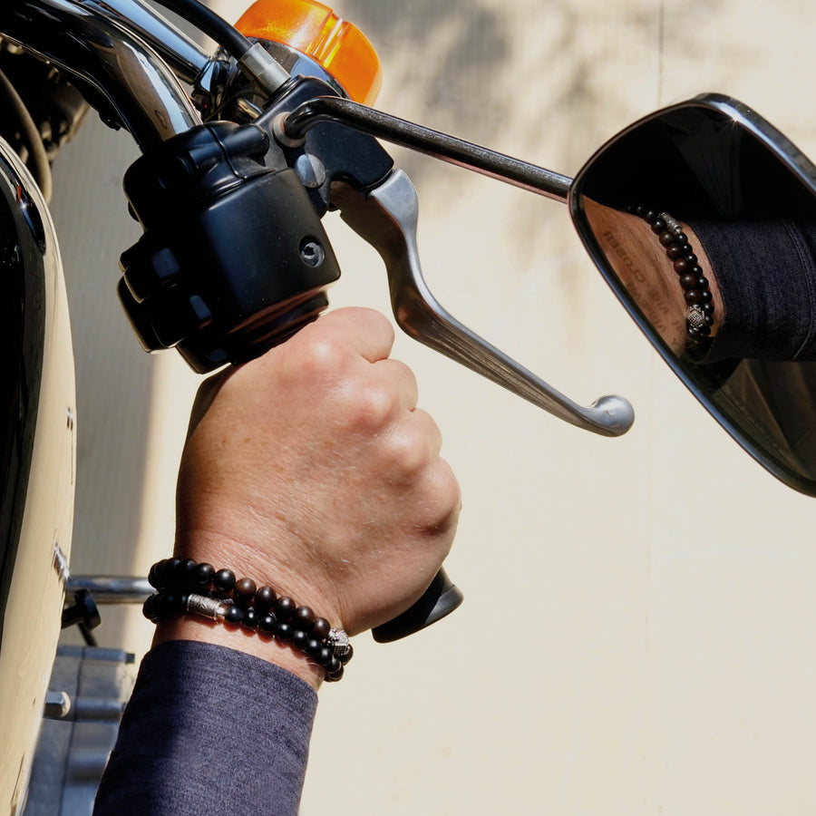 Close view of male model's wrist on a Harley Davidson Bike wearing the ALEC Men beaded bracelet made with Ebony and a sterling silver center piece and the MAXIM Men beaded bracelet made with Mat Black Onyx and sterling silver elements. 
