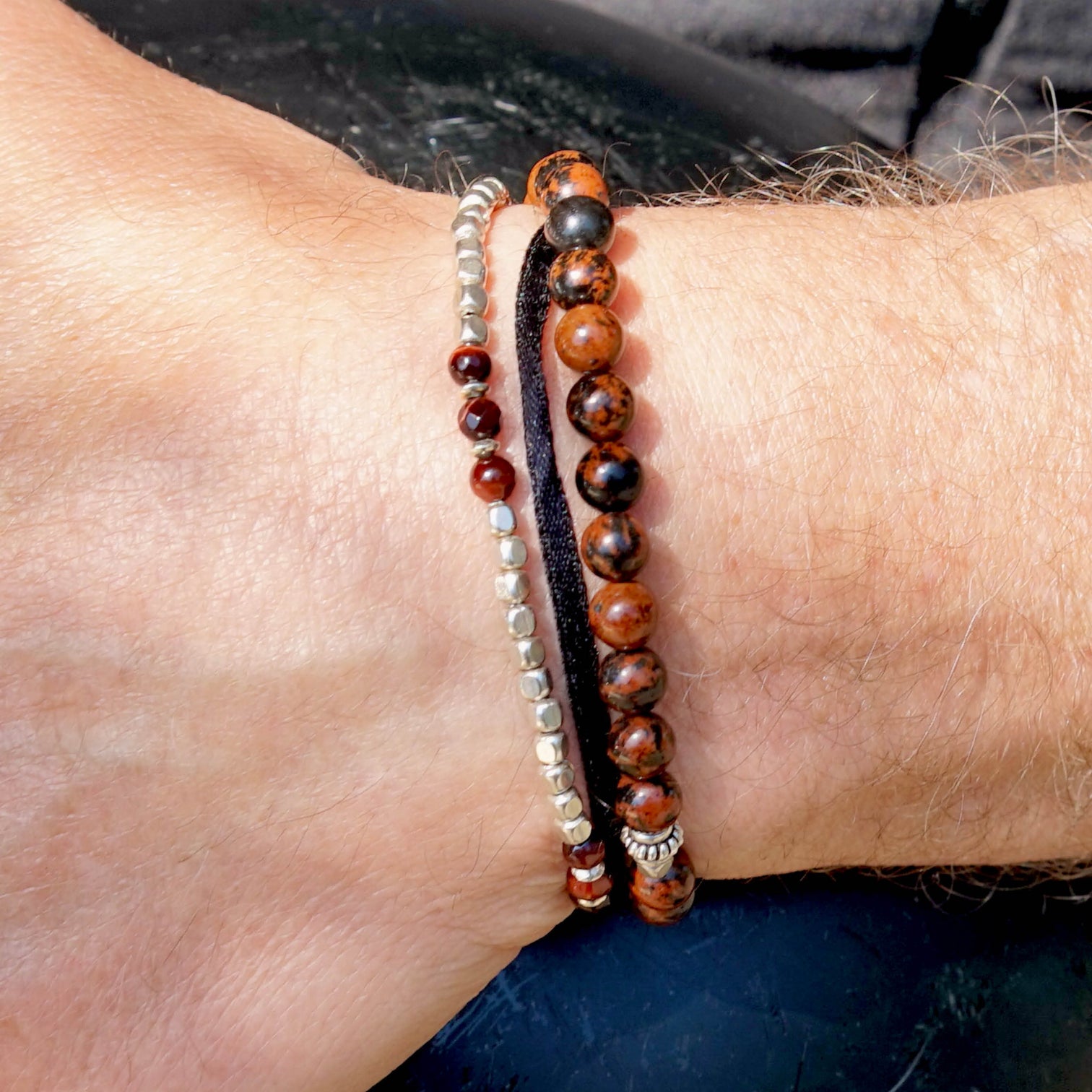 Close-up of a male model is wearing two Men Beaded Bracelets. One of the bracelet is the ARIEL Bracelet made with Red Tiger Eye and Sterling Silver Elements. The other bracelet is the KANE Bracelet made with Mahogany Obsidian and Sterling Silver Elements.