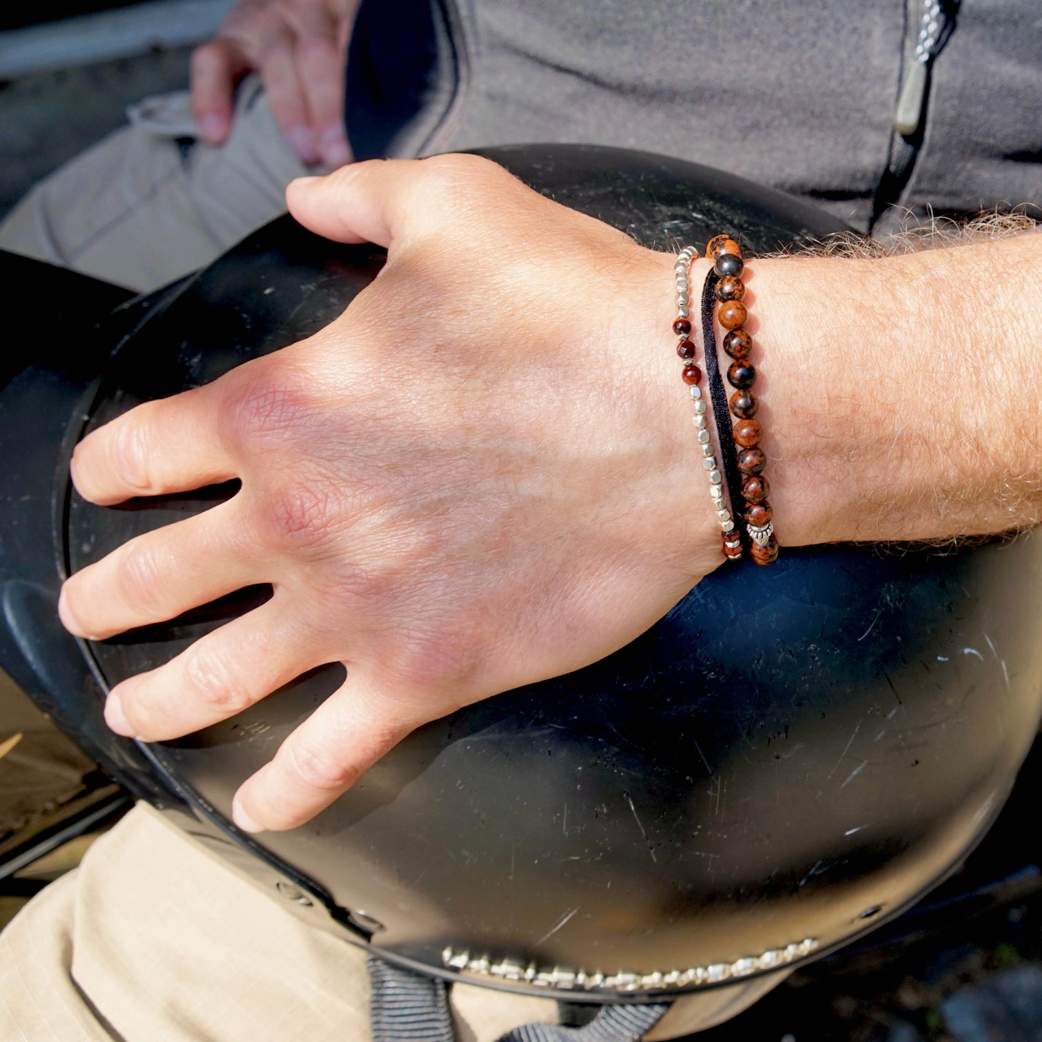 A male model is wearing two Men Beaded Bracelets. One of the bracelet is the ARIEL Bracelet made with Red Tiger Eye and Sterling Silver Elements. The other bracelet is the KANE Bracelet made with Mahogany Obsidian and Sterling Silver Elements.