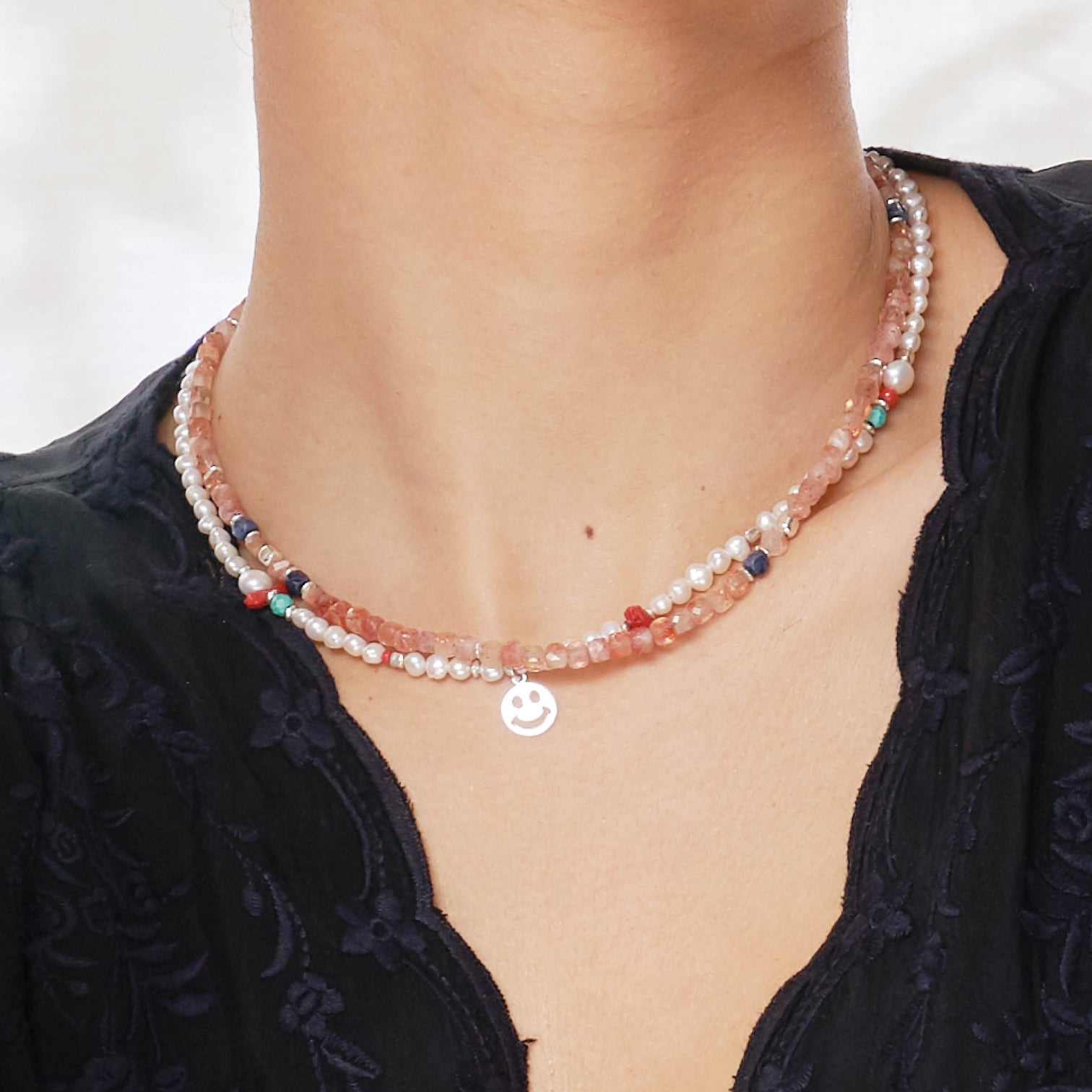 Close-Up of a female model wearing the Ginger Necklace made with faceted square Sunstones, and the Alaia Necklace strung with Freshwater White Pearls and a Smiley charm. 